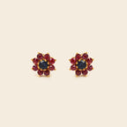 A pair of gold flower-shaped stud earrings featuring a dark blue center stone surrounded by red gemstones, set against a solid cream background.