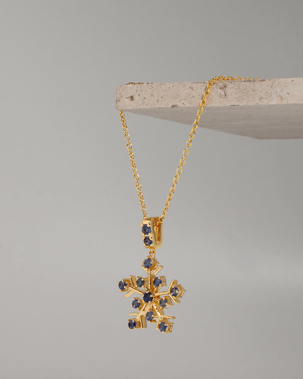 A gold snowflake pendant necklace with small dark blue gemstone accents, hanging from a gold chain draped over a light stone display block against a neutral gray background.