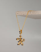 A gold snowflake pendant necklace with small dark blue gemstone accents, hanging from a gold chain draped over a light stone display block against a neutral gray background.