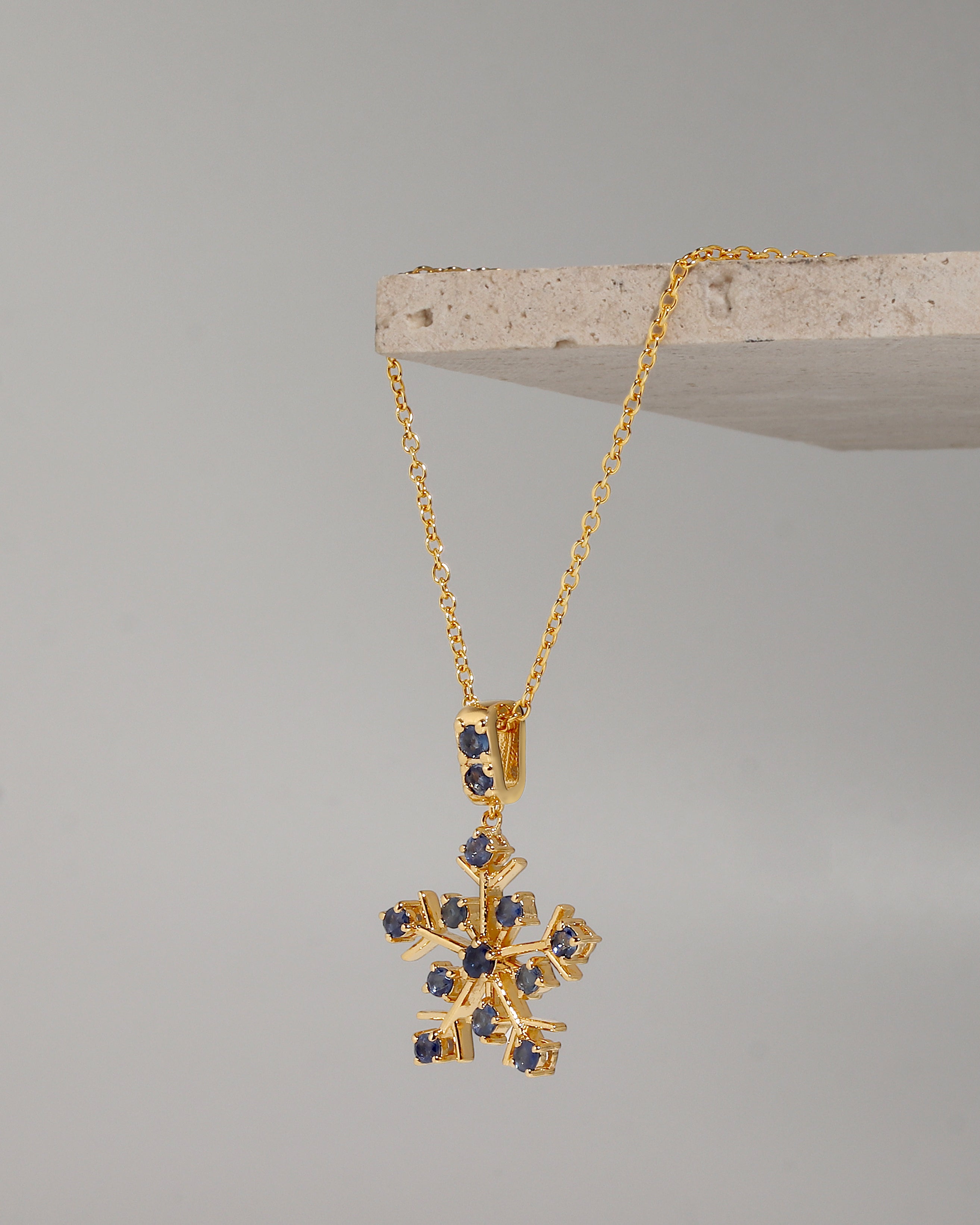 A gold snowflake pendant necklace with small dark blue gemstone accents, hanging from a gold chain draped over a light stone display block against a neutral gray background.