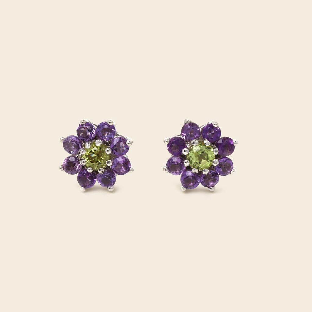 A pair of flower-shaped stud earrings featuring a light green peridot center surrounded by a halo of round purple amethyst petals in silver-toned metal, set against a solid light beige background.