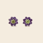 A pair of flower-shaped stud earrings featuring a light green peridot center surrounded by a halo of round purple amethyst petals in silver-toned metal, set against a solid light beige background.
