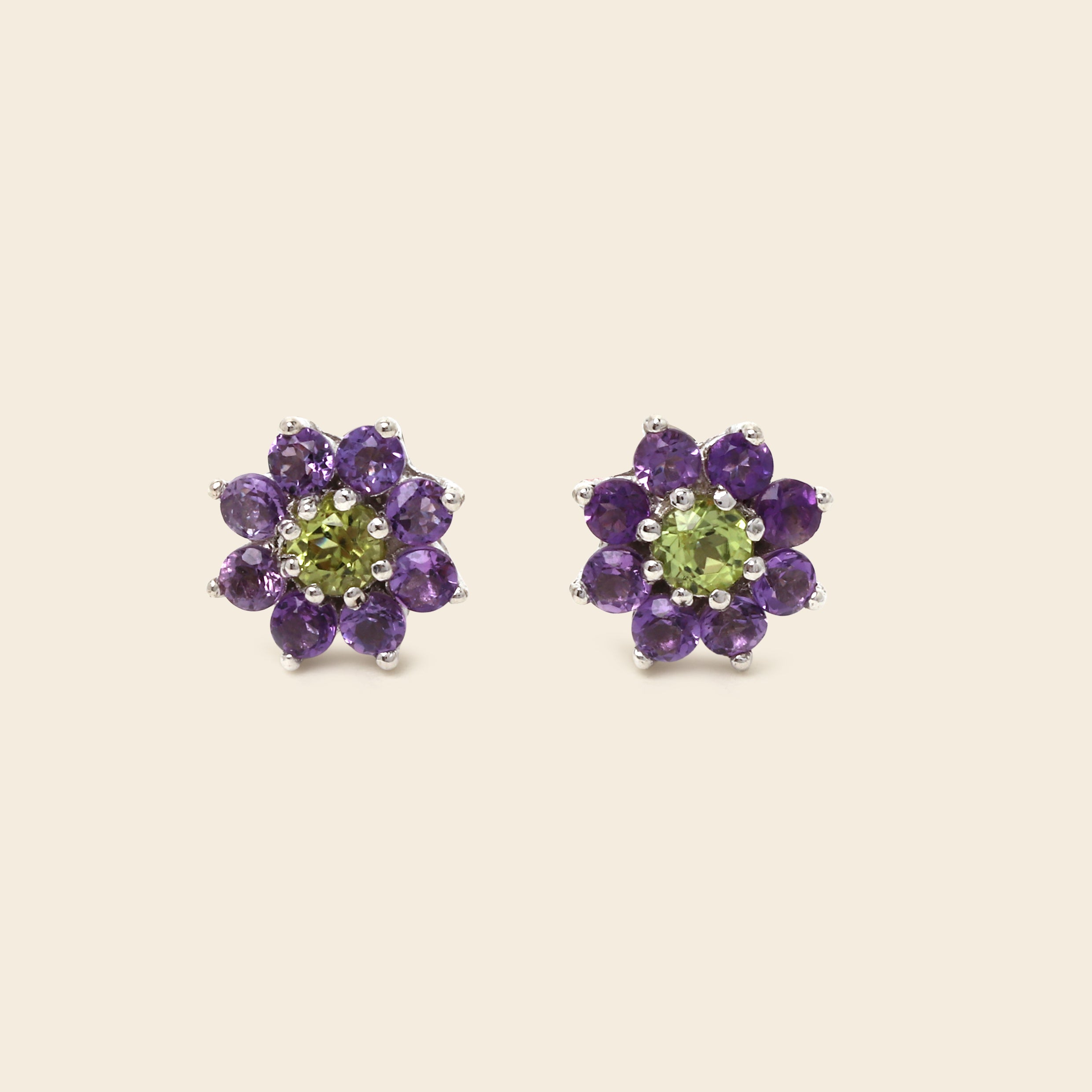 A pair of flower-shaped stud earrings featuring a light green peridot center surrounded by a halo of round purple amethyst petals in silver-toned metal, set against a solid light beige background.