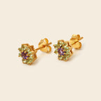 A pair of gold flower stud earrings featuring a central purple amethyst and surrounded by light green peridot gemstones, displayed on a warm cream background.