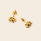 A pair of gold stud earrings with small green gemstone clusters and a central purple gemstone, shown on a light beige background.