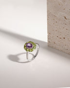 A silver ring featuring a round purple amethyst center stone surrounded by a halo of green peridot gemstones, with a pavé band of small clear stones, photographed on a white surface next to a textured stone block.