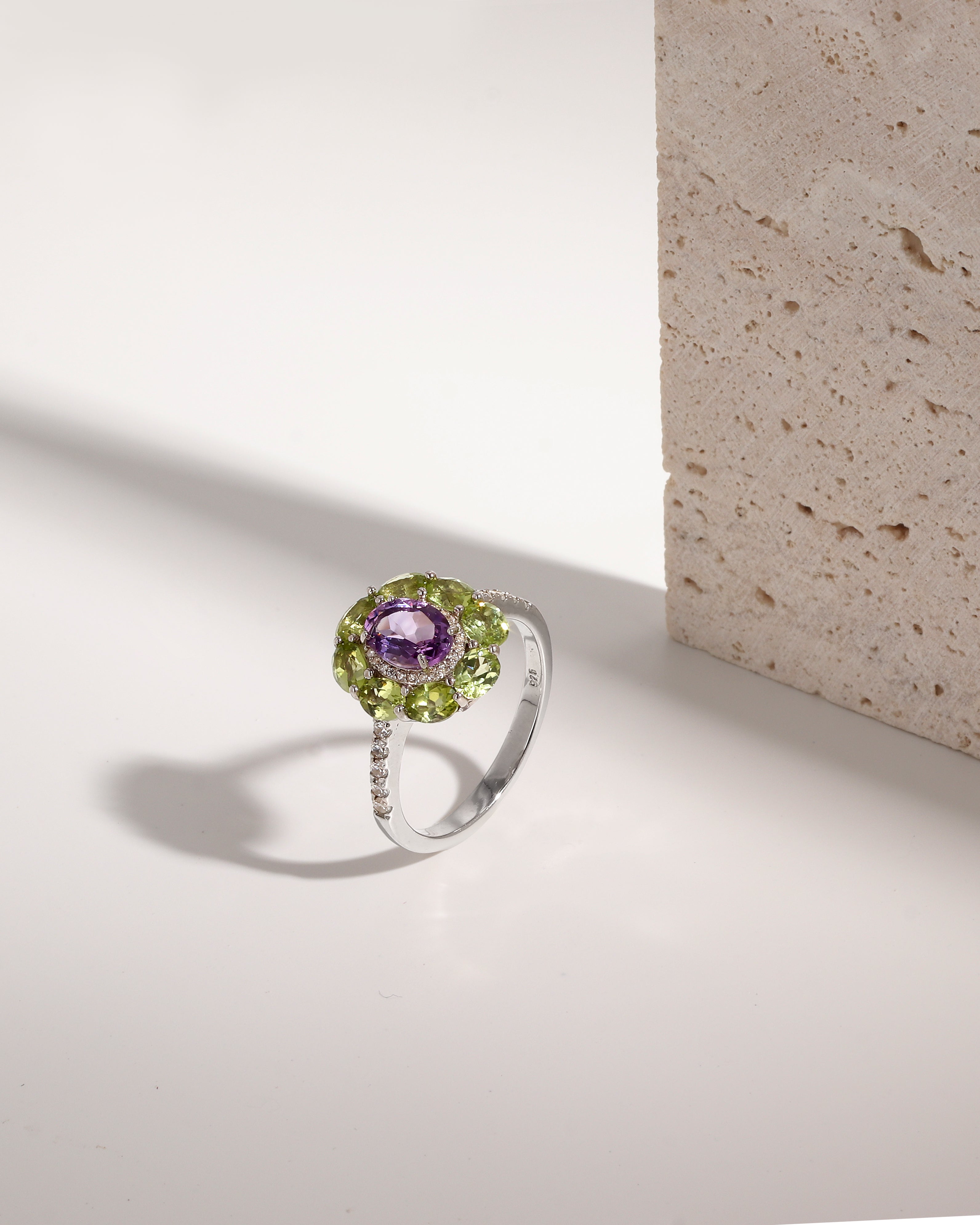A silver ring featuring a round purple amethyst center stone surrounded by a halo of green peridot gemstones, with a pavé band of small clear stones, photographed on a white surface next to a textured stone block.