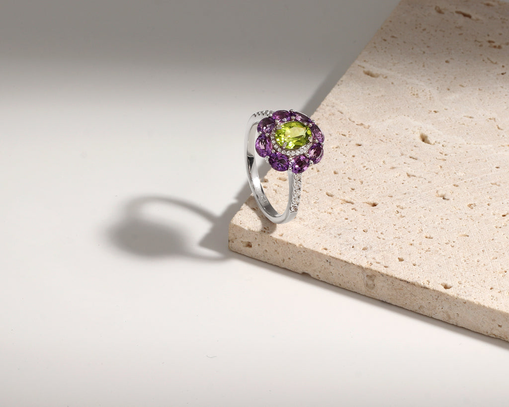 A silver ring featuring a central green peridot gemstone surrounded by a halo of purple amethyst stones, displayed on a textured beige stone slab against a clean white background.