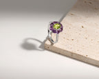 A silver ring featuring a central green peridot gemstone surrounded by a halo of purple amethyst stones, displayed on a textured beige stone slab against a clean white background.