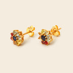 A pair of gold stud earrings with a multi-colored gemstone cluster, featuring a dark blue center stone surrounded by yellow, light blue, and red stones, set against a solid cream background.