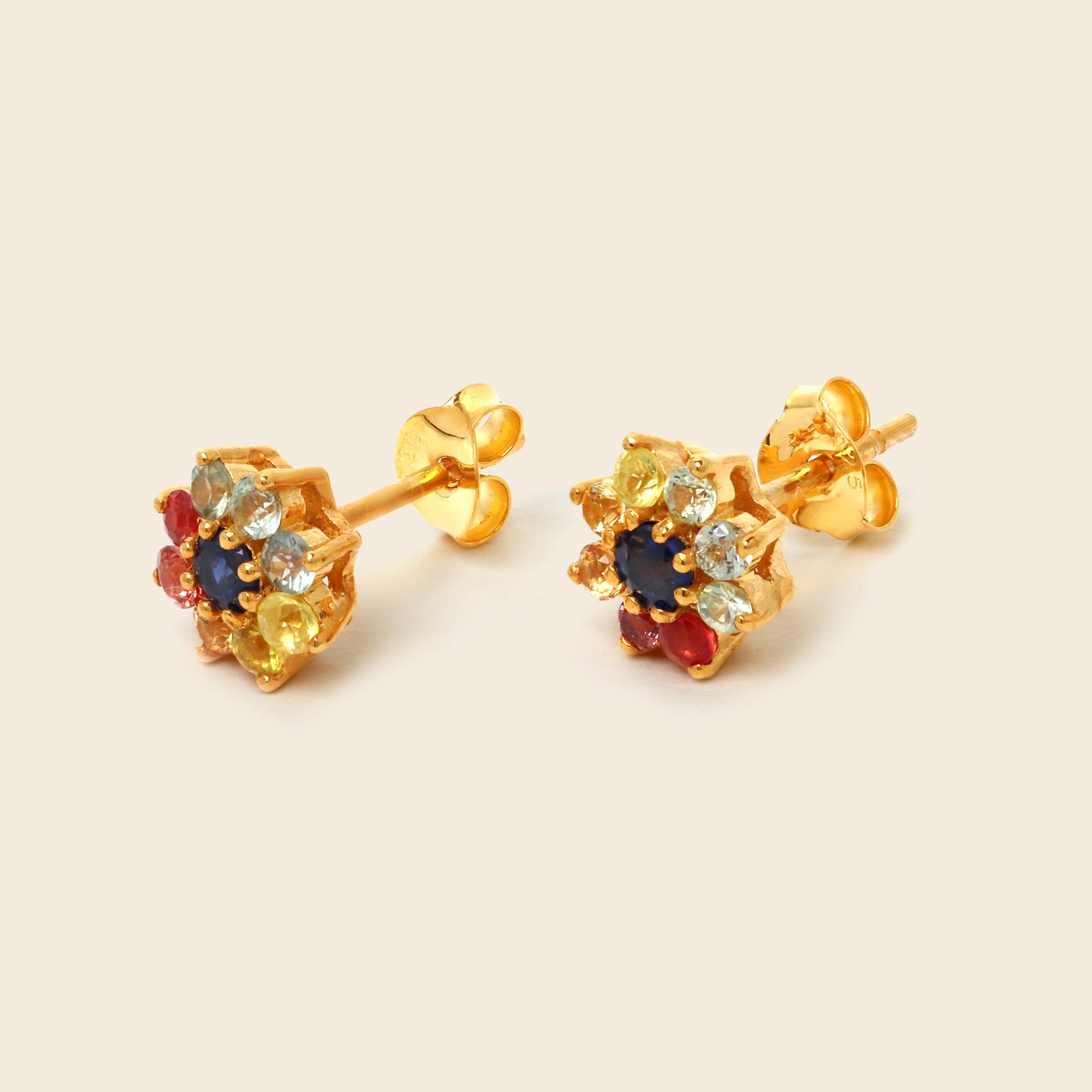 A pair of gold stud earrings with a multi-colored gemstone cluster, featuring a dark blue center stone surrounded by yellow, light blue, and red stones, set against a solid cream background.