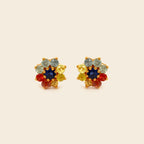 A pair of gold-toned stud earrings featuring a multi-colored floral design with a dark blue center stone, surrounded by yellow, light blue, and red stones, set against a solid cream background.