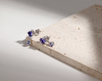 A pair of silver stud earrings with round blue gemstones, resting on a textured beige stone slab against a white background with dramatic shadows.