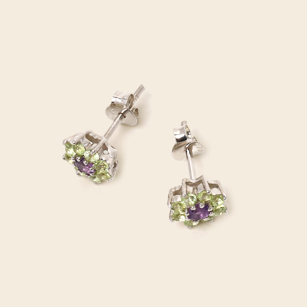 A pair of silver stud earrings with a central purple amethyst and a surrounding halo of light green peridot gemstones, displayed on a light beige background.