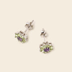 A pair of silver stud earrings with a central purple amethyst and a surrounding halo of light green peridot gemstones, displayed on a light beige background.