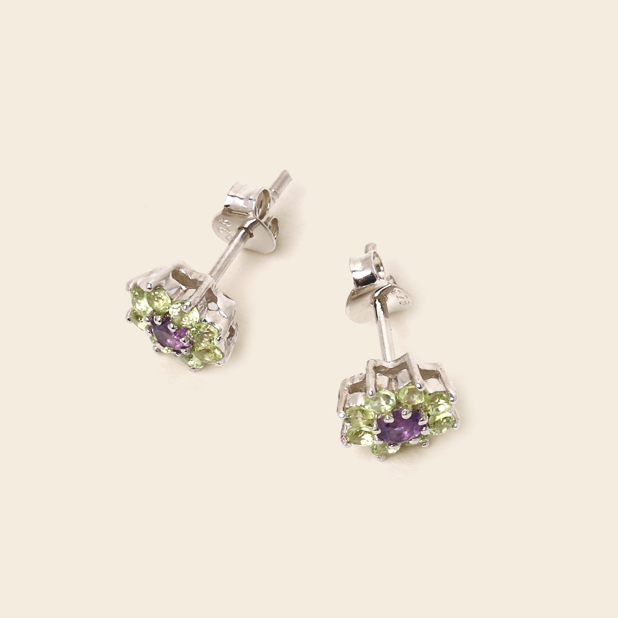 A pair of silver stud earrings with a central purple amethyst and a surrounding halo of light green peridot gemstones, displayed on a light beige background.