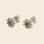 A pair of silver stud earrings featuring a central round purple gemstone surrounded by a halo of small green gemstones, shown on a light beige background.