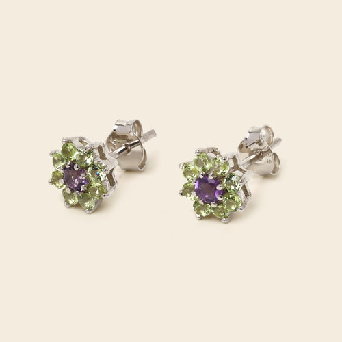A pair of silver stud earrings featuring a central round purple gemstone surrounded by a halo of small green gemstones, shown on a light beige background.