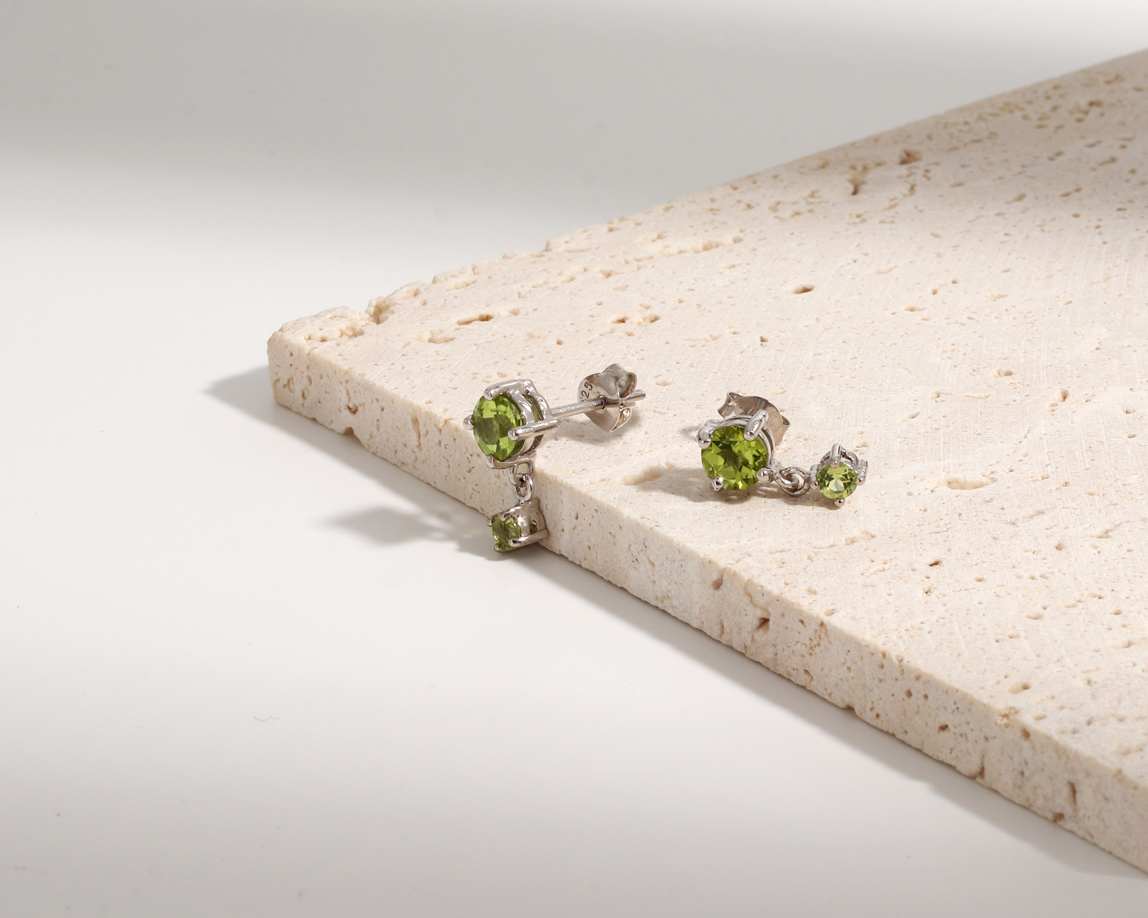 A pair of silver stud earrings with green gemstones, displayed on a light beige stone slab against a clean white background.