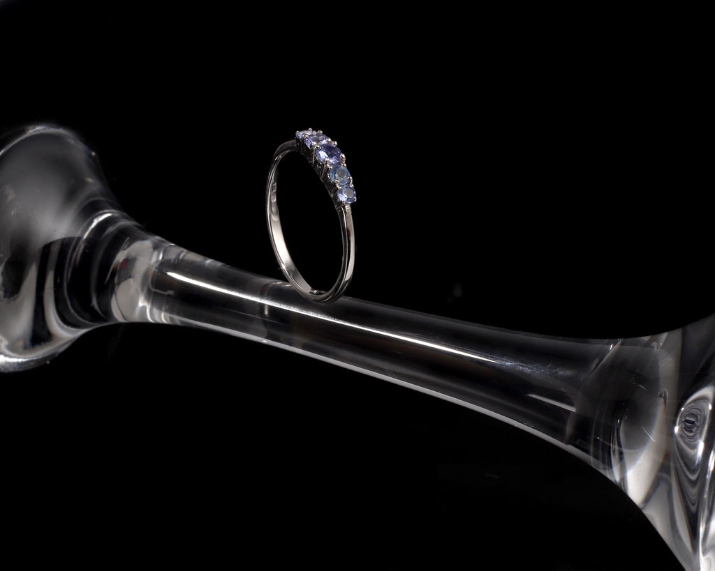 A silver ring with a row of small clear stones, displayed on the stem of a clear glass goblet against a solid black background.