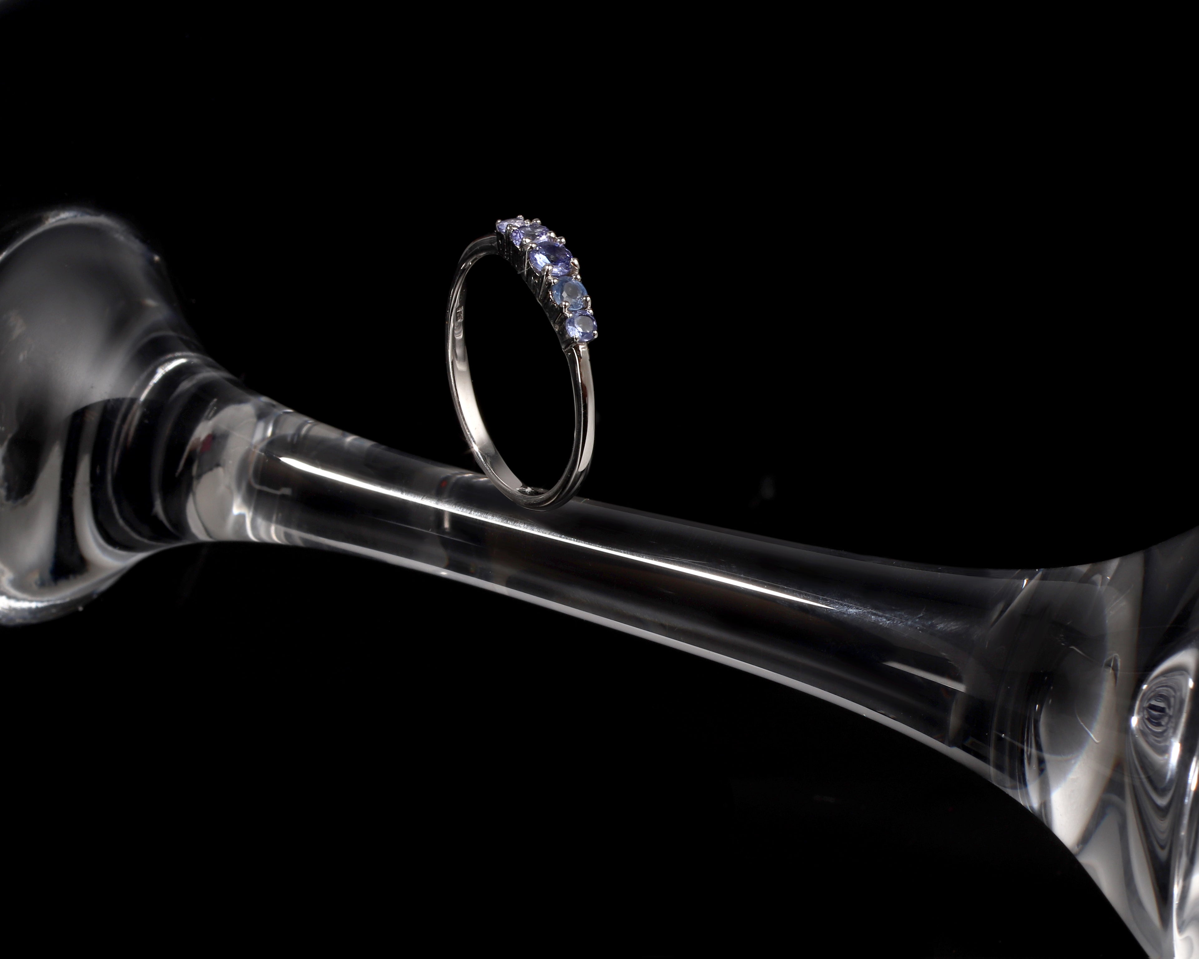 A silver ring with a row of small clear stones, displayed on the stem of a clear glass goblet against a solid black background.