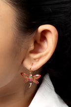 A close-up of a person's ear wearing a dangling dragonfly earring. The earring is gold-toned with red and pink enamel wings and a small green center detail.