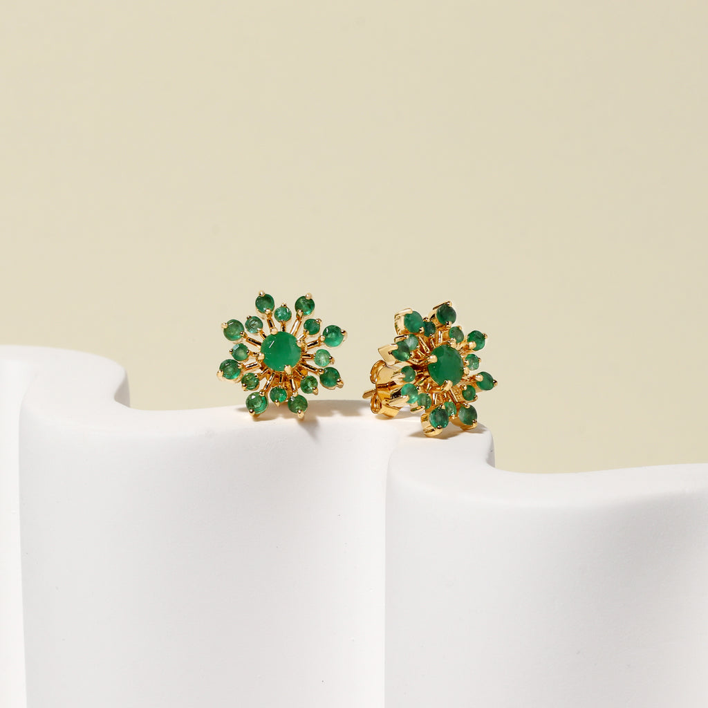A pair of gold flower-shaped stud earrings with green gemstones, displayed on a white sculptural stand against a light beige background.