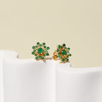 A pair of gold flower-shaped stud earrings with green gemstones, displayed on a white sculptural stand against a light beige background.