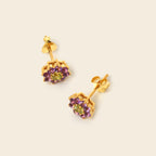 A pair of gold stud earrings featuring a central green gemstone surrounded by small purple gemstones in a geometric setting, displayed on a light beige background.