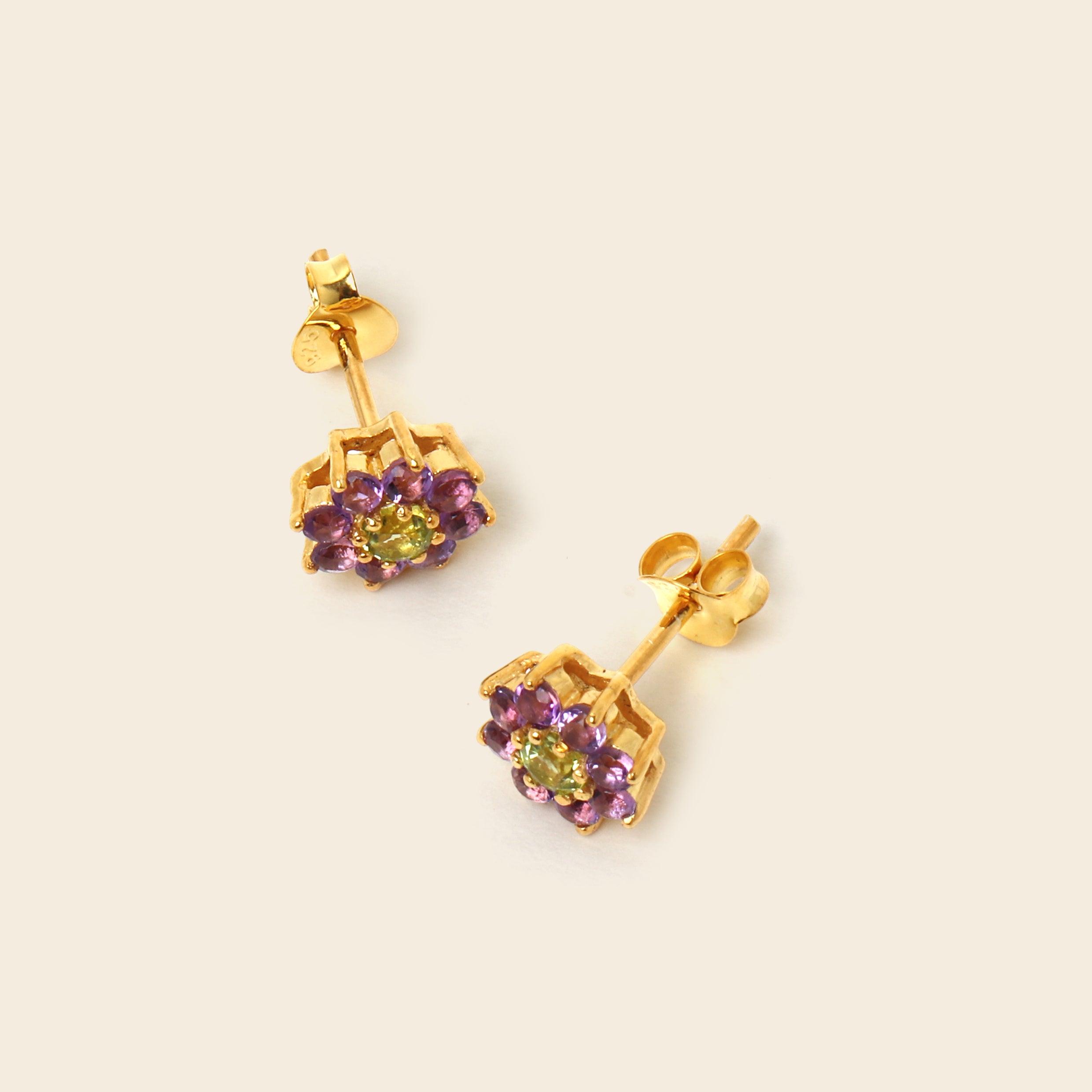 A pair of gold stud earrings featuring a central green gemstone surrounded by small purple gemstones in a geometric setting, displayed on a light beige background.