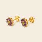 A pair of gold flower stud earrings with purple gemstone petals and a light green center stone, shown on a solid light beige background.