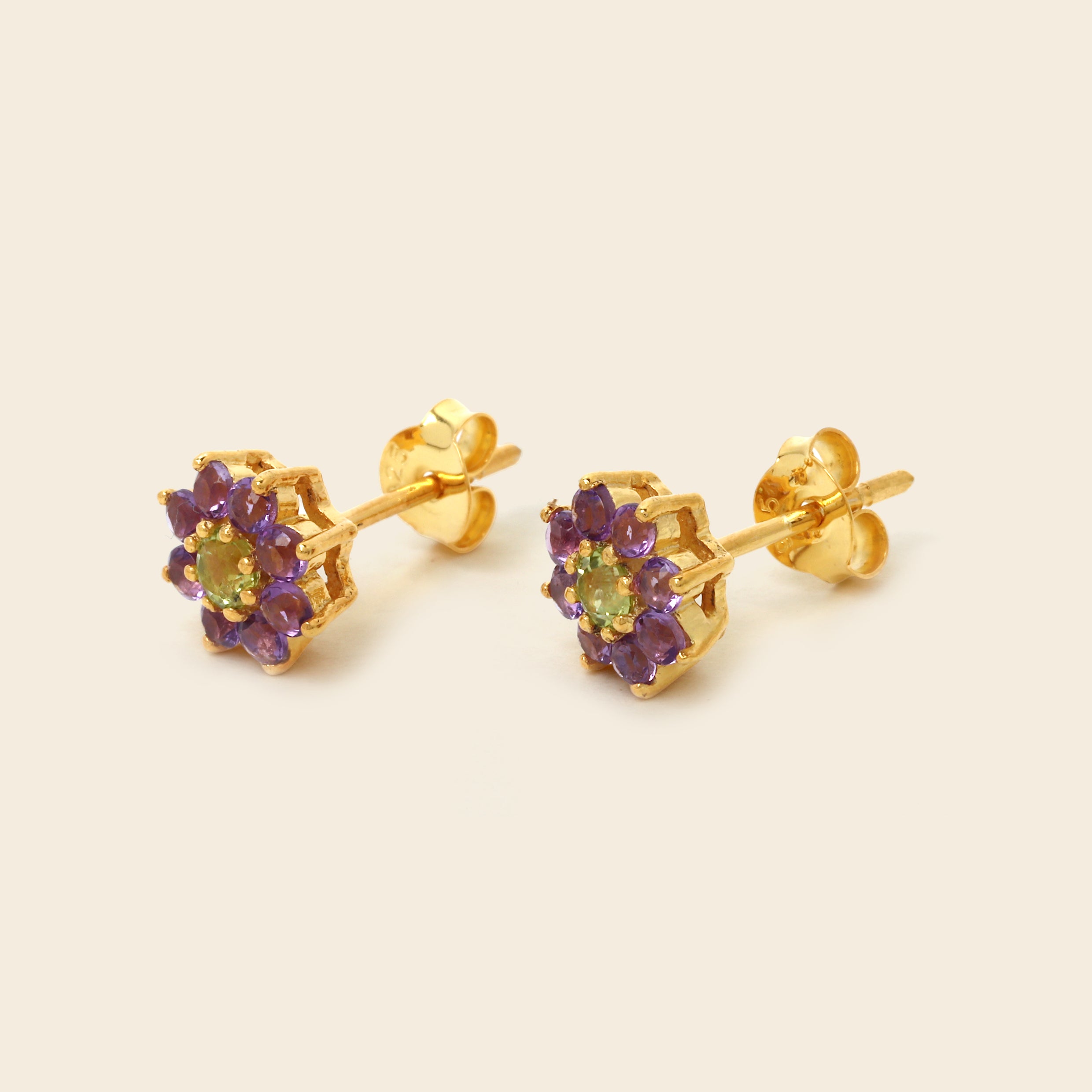 A pair of gold flower stud earrings with purple gemstone petals and a light green center stone, shown on a solid light beige background.
