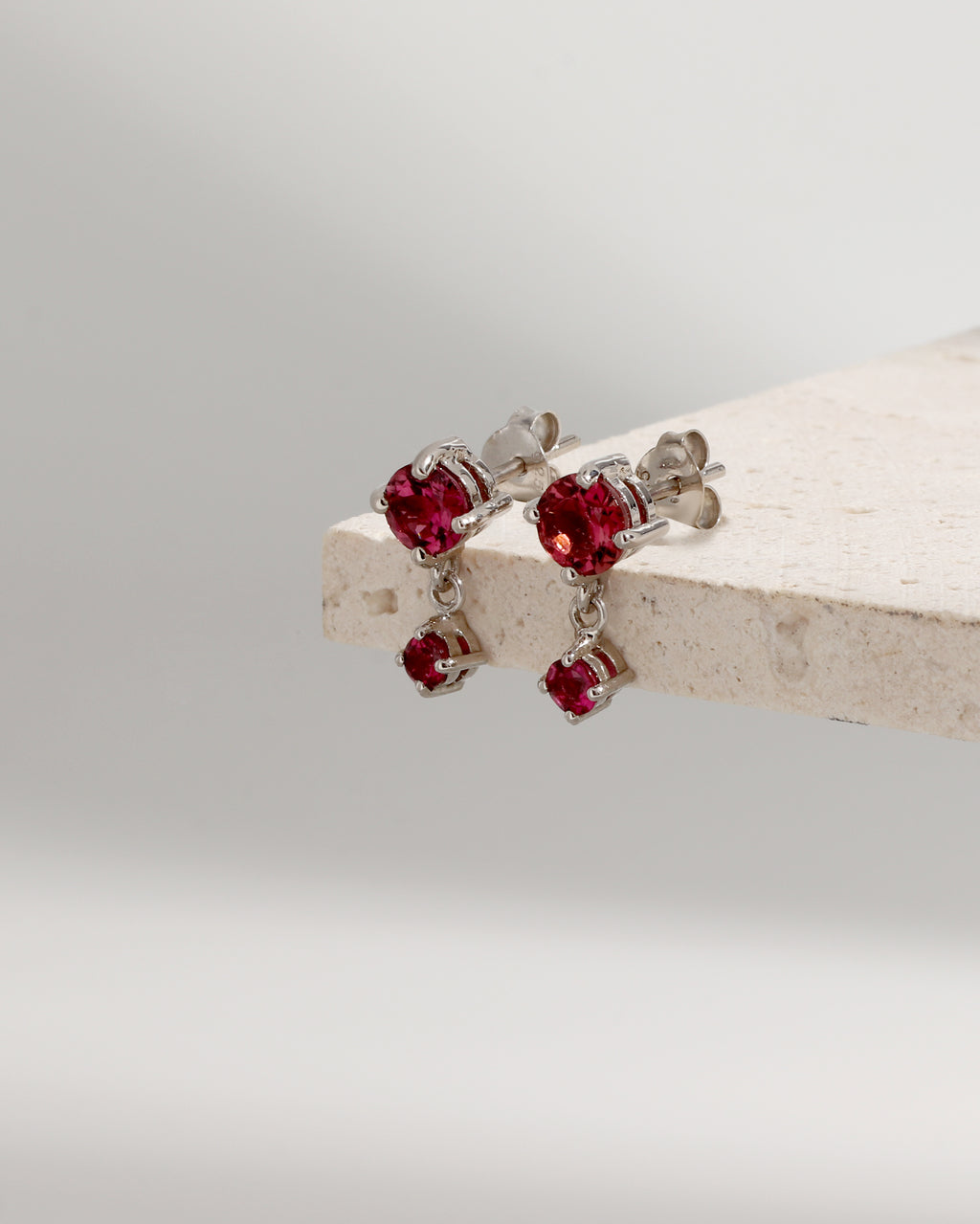 A pair of silver stud earrings with dangling red gemstone charms, displayed on a light beige stone block against a soft gray background.