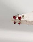 A pair of silver stud earrings with dangling red gemstone charms, displayed on a light beige stone block against a soft gray background.