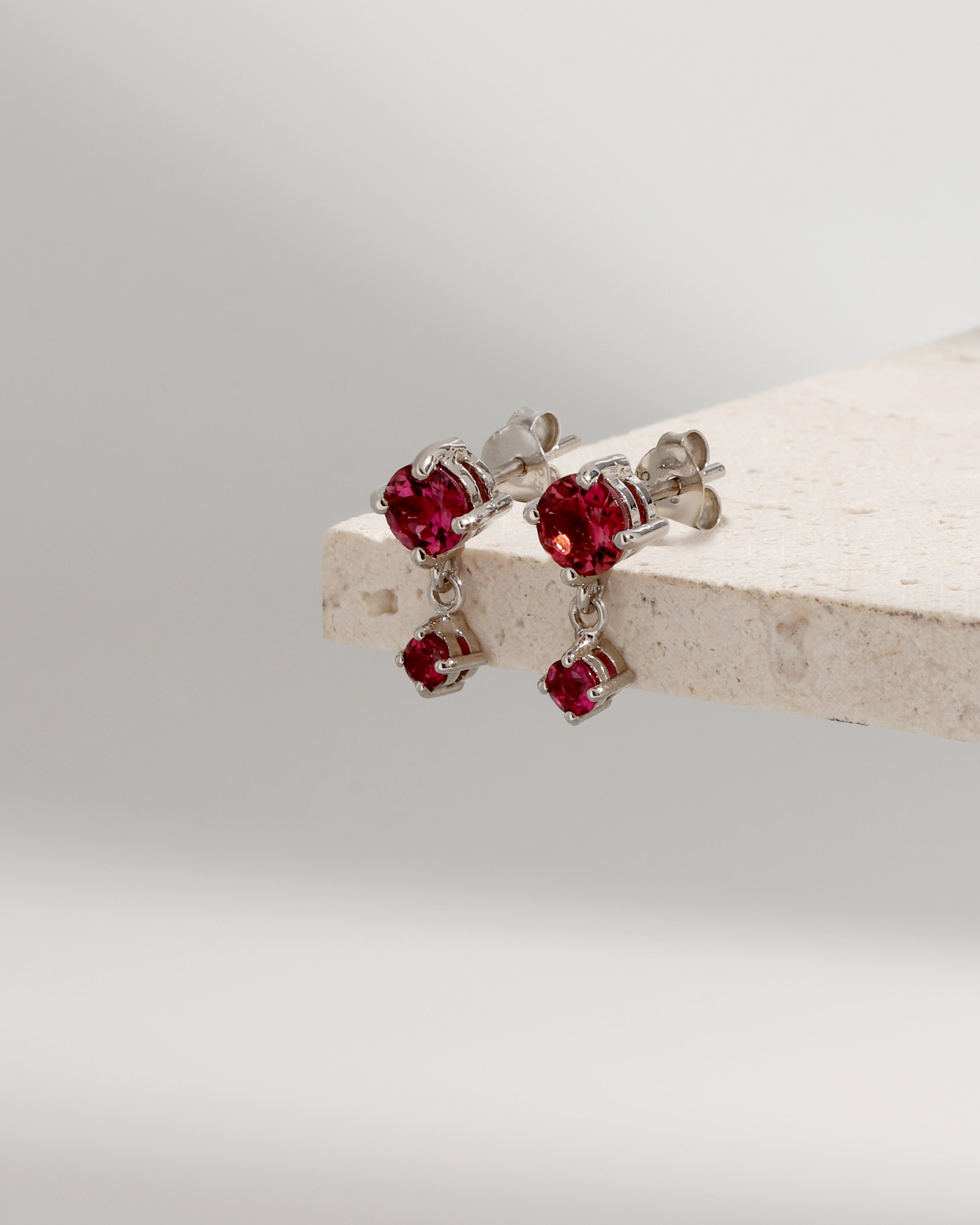 A pair of silver stud earrings with dangling red gemstone charms, displayed on a light beige stone block against a soft gray background.