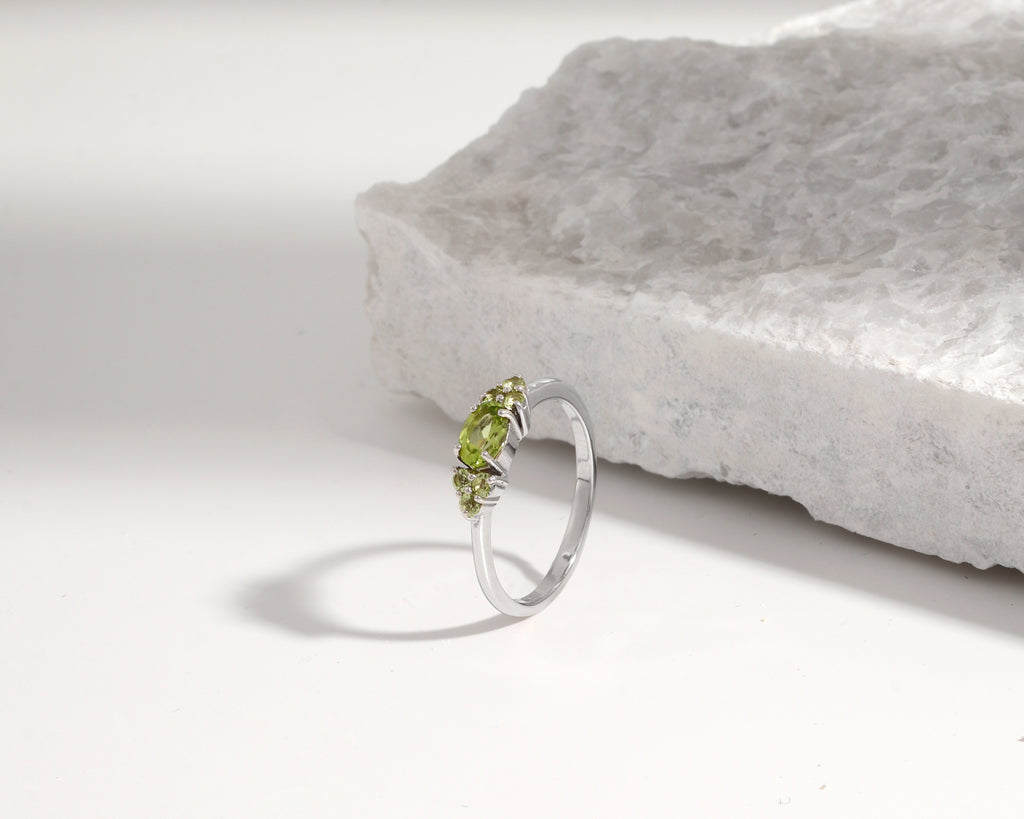 A silver ring with three small green gemstones, photographed on a white surface next to a textured white stone slab.