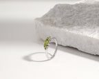 A silver ring with three small green gemstones, photographed on a white surface next to a textured white stone slab.