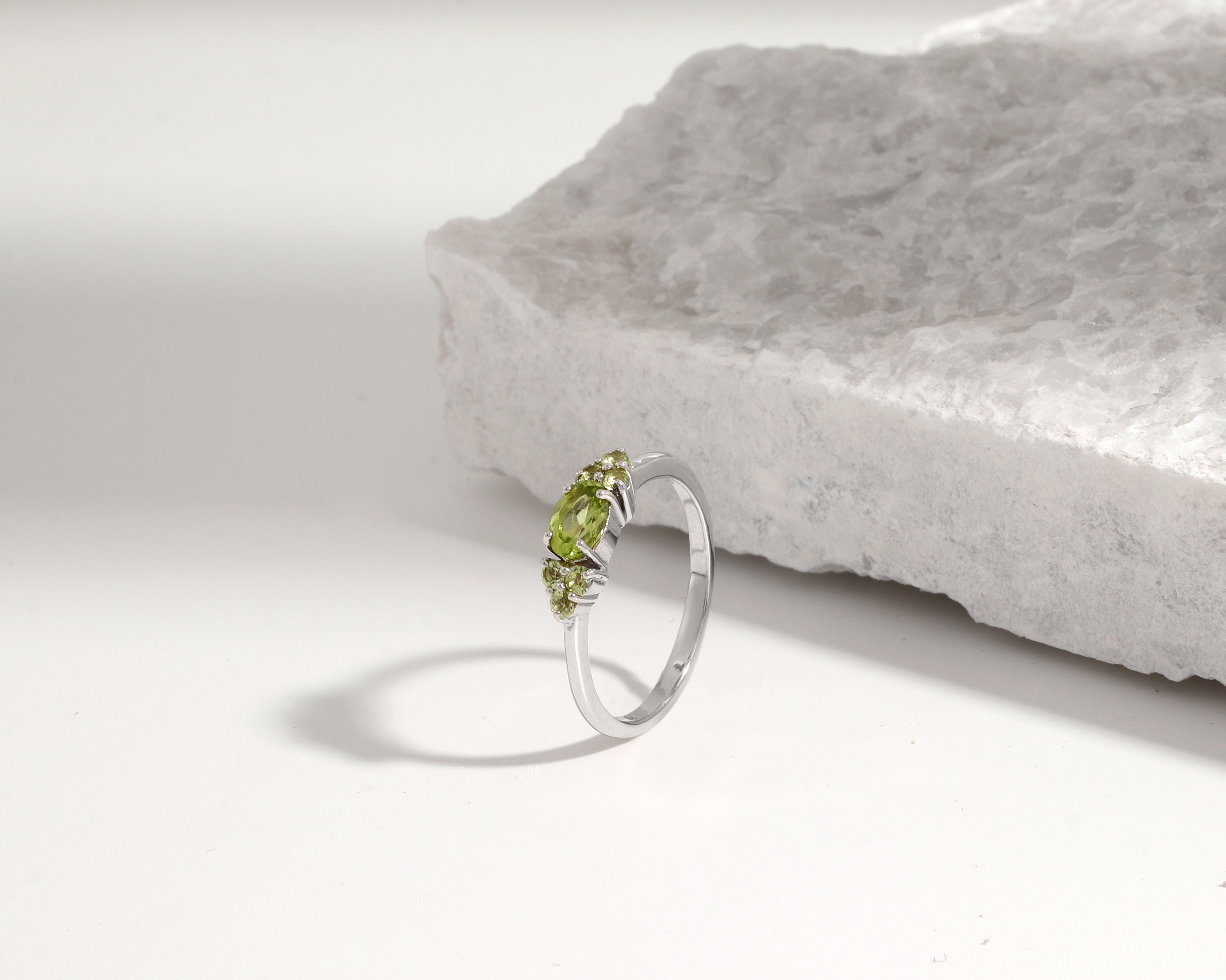 A silver ring with three small green gemstones, photographed on a white surface next to a textured white stone slab.