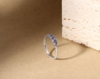 A silver band ring featuring a curved row of deep blue gemstones, photographed on a textured beige surface next to a light stone prop.