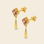 A pair of gold drop earrings featuring a square cluster of small pinkish-purple gemstones at the top and a teardrop-shaped gold drop hanging below, shown on a light beige background.