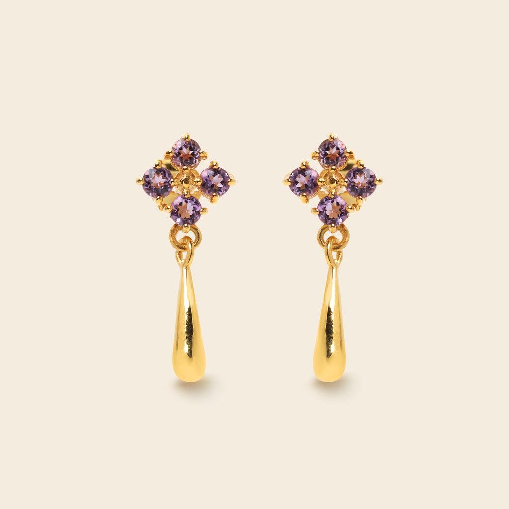 A pair of gold dangle earrings with a small cluster of purple gemstones at the top and a polished gold teardrop drop below, shown on a light beige background.