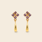 A pair of gold dangle earrings with a small cluster of purple gemstones at the top and a polished gold teardrop drop below, shown on a light beige background.