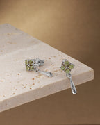 A pair of silver drop earrings with green gemstone clusters, displayed on a light beige stone slab against a warm brown background.