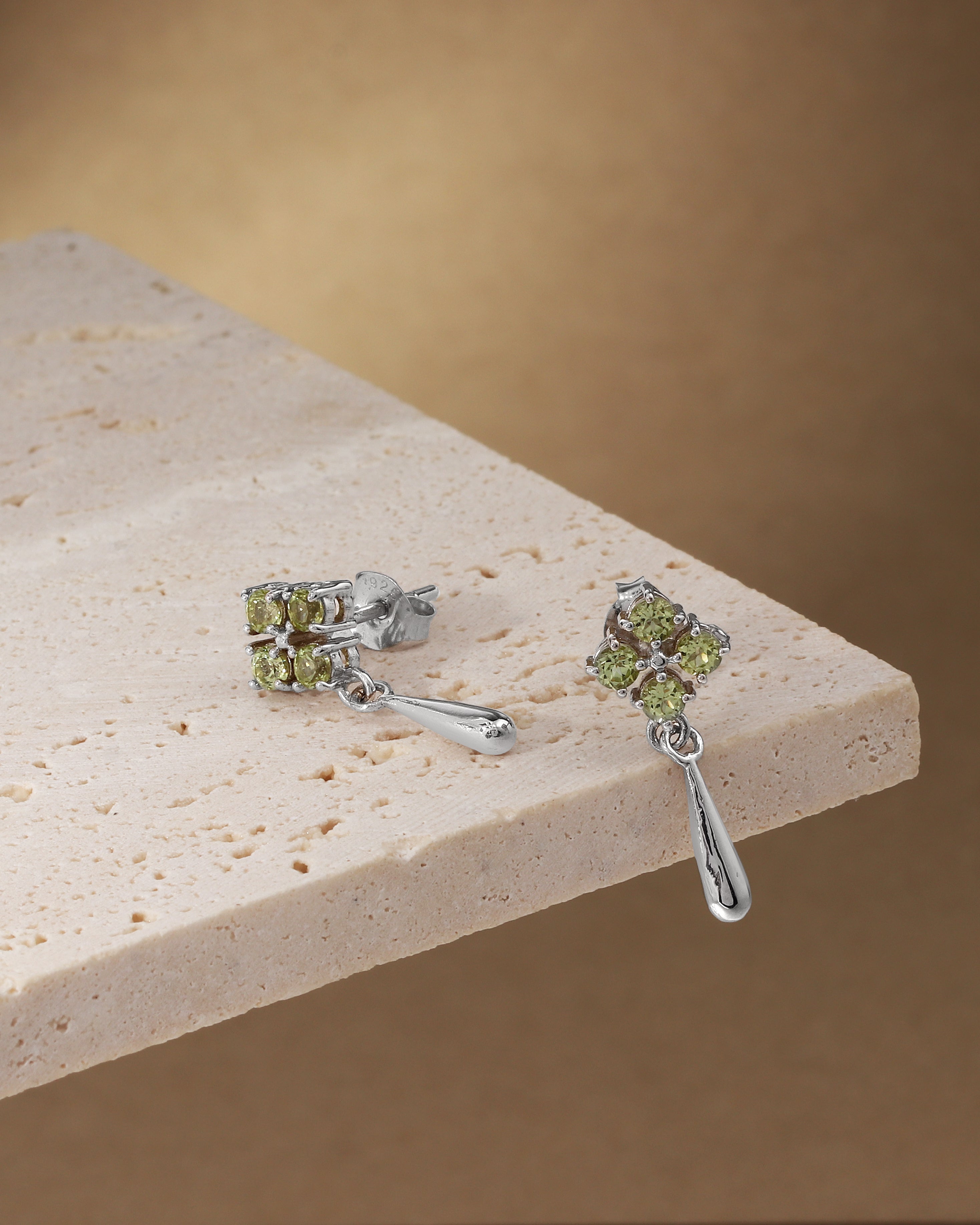 A pair of silver drop earrings with green gemstone clusters, displayed on a light beige stone slab against a warm brown background.