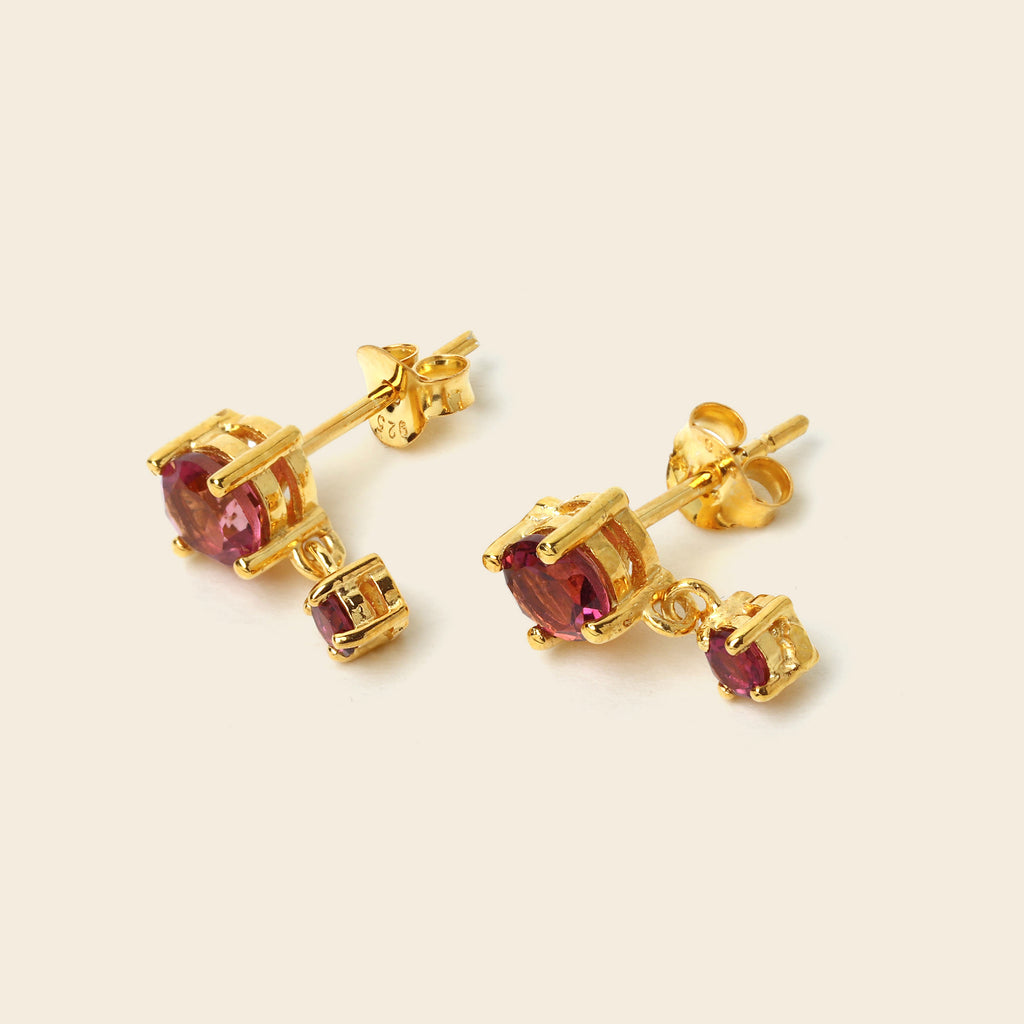 A pair of gold stud earrings with oval pinkish-purple gemstones and smaller rectangular accent stones, shown on a light beige background.