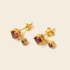 A pair of gold stud earrings with oval pinkish-purple gemstones and smaller rectangular accent stones, shown on a light beige background.