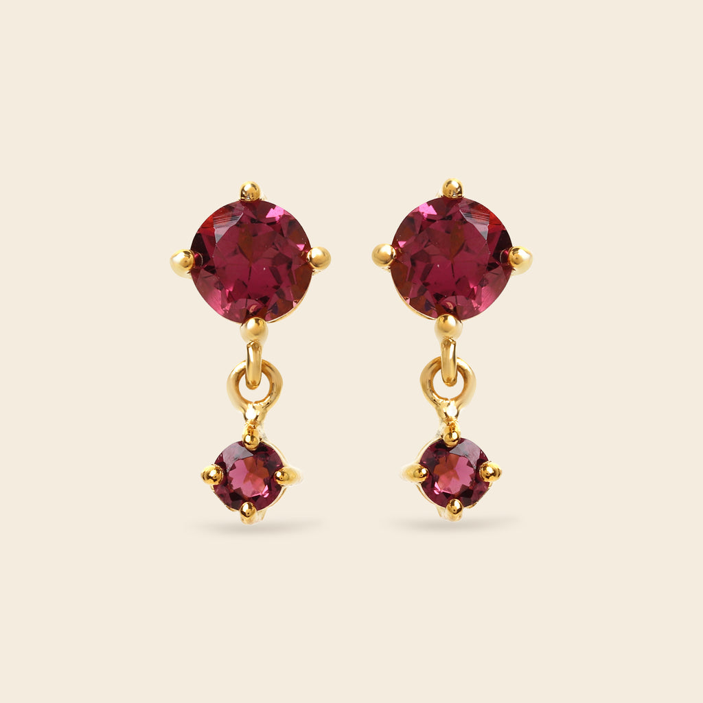 A pair of gold drop earrings featuring round deep pinkish-red gemstones, with a larger round stone at the top and a smaller teardrop-shaped stone dangling below, set against a solid light beige background.