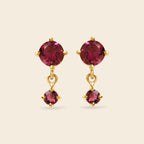 A pair of gold drop earrings featuring round deep pinkish-red gemstones, with a larger round stone at the top and a smaller teardrop-shaped stone dangling below, set against a solid light beige background.