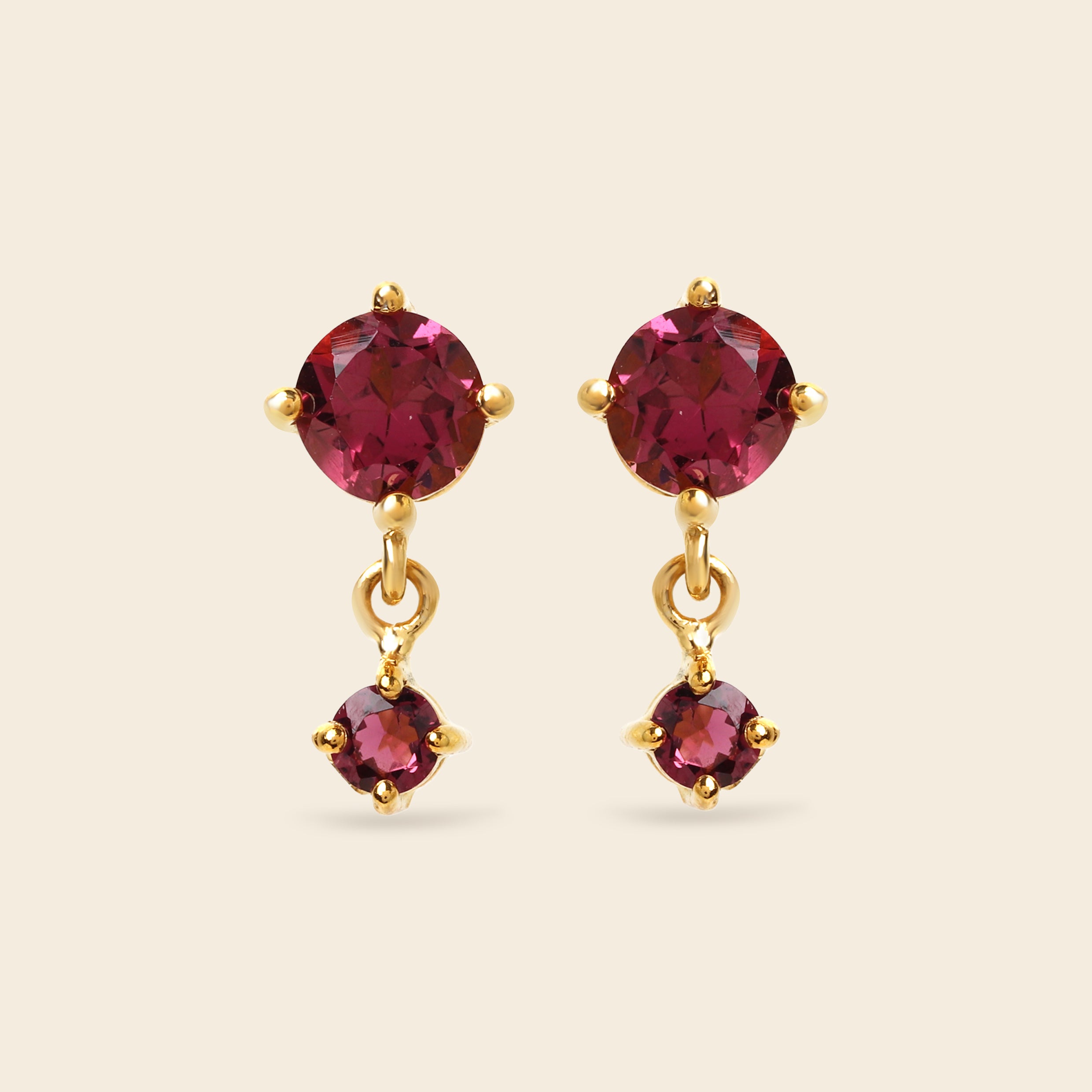 A pair of gold drop earrings featuring round deep pinkish-red gemstones, with a larger round stone at the top and a smaller teardrop-shaped stone dangling below, set against a solid light beige background.