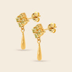 A pair of gold dangle earrings featuring a cluster of round-cut light green gemstones in a floral setting, with a polished gold teardrop drop hanging below, shown on a light beige background.